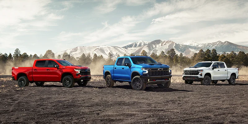 The image features three Chevrolet Silverado trucks in different colors—red, blue, and white—parked on rugged terrain with snow-capped mountains in the background. The trucks are showcased in a dynamic, off-road setting, highlighting their powerful design.
