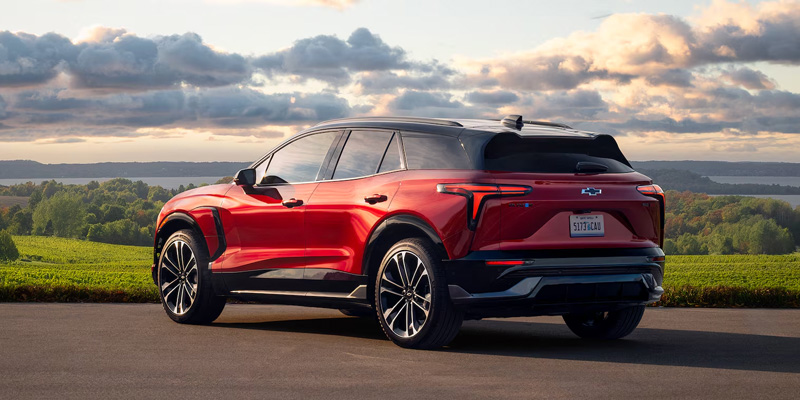 A sleek, red Blazer EV with a modern design is parked on a country road. The background features lush green fields and a sky with dramatic clouds.