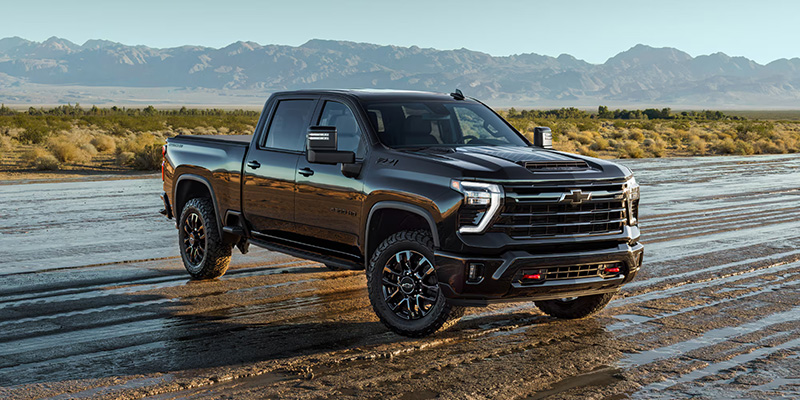 A black pickup truck Silverado HD parked on muddy terrain, with mountains in the background under a clear blue sky, conveying strength and adventure.